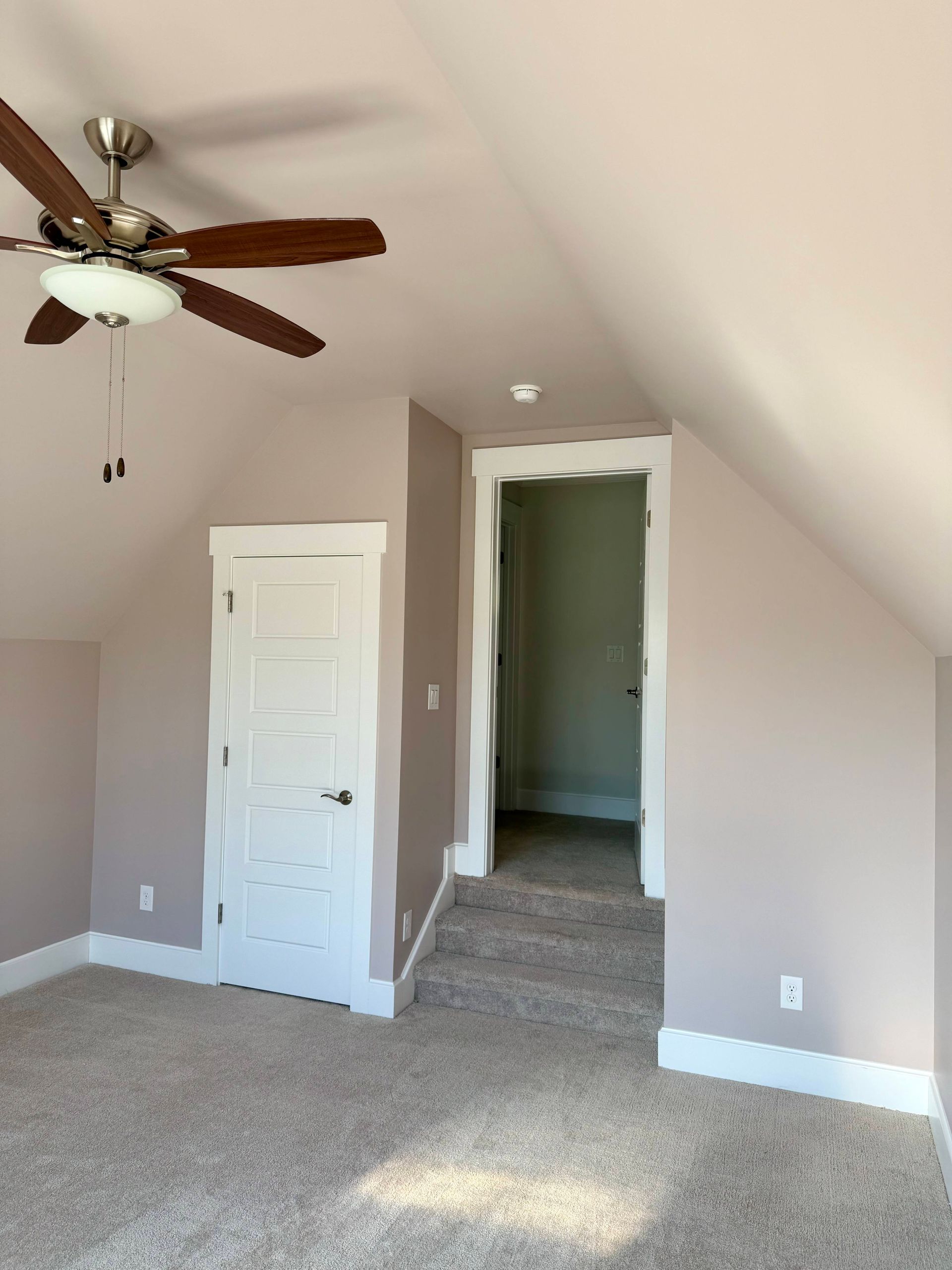 Empty carpeted bedroom with sloped ceiling, ceiling fan, white door, and doorway to a hall