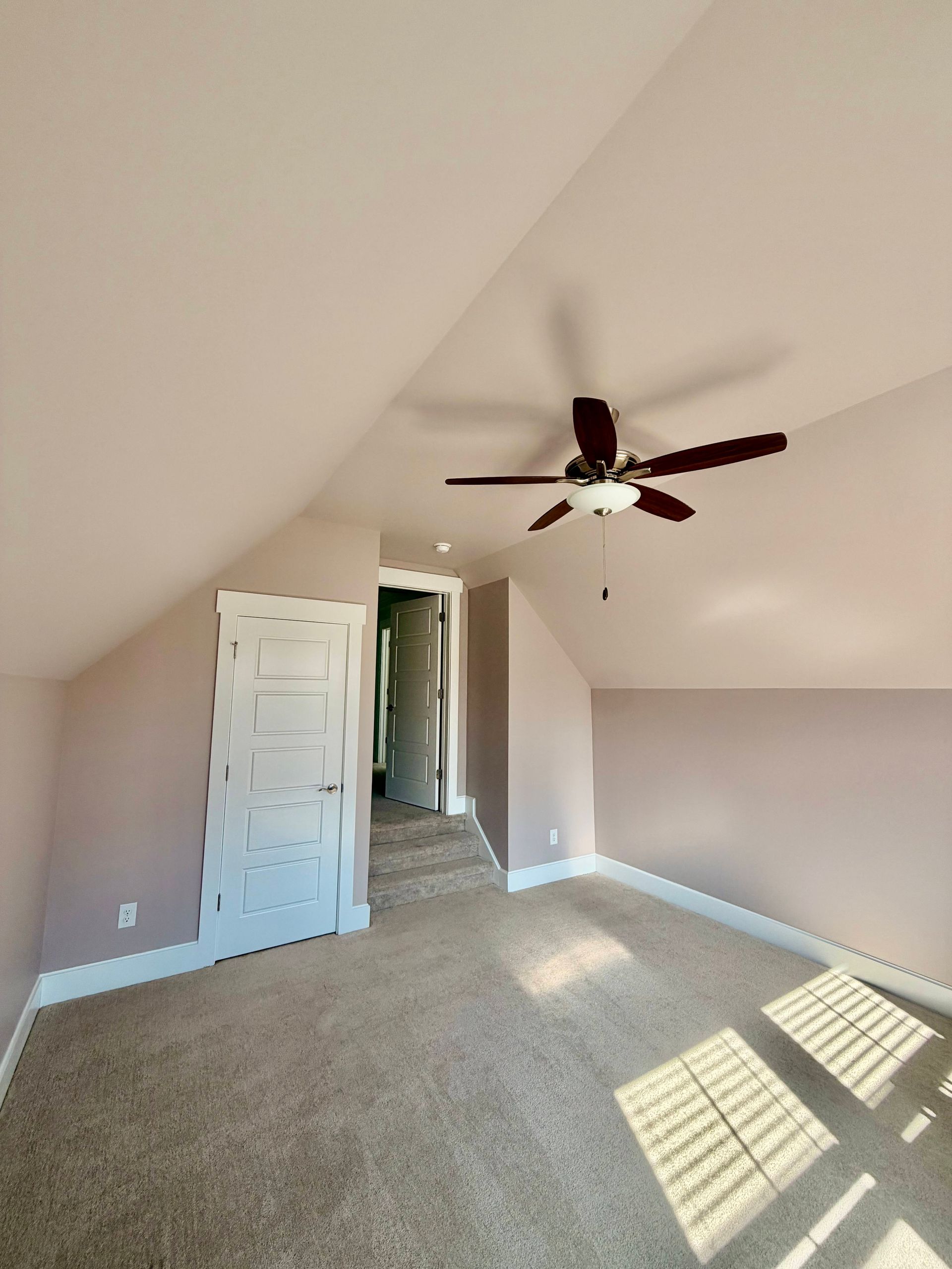 Empty carpeted bedroom with sloped ceiling, open door, ceiling fan, and sunlight on the floor.