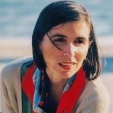 Donna all'aperto in riva all'acqua, con indosso una sciarpa rossa e un maglione beige, che guarda di lato.