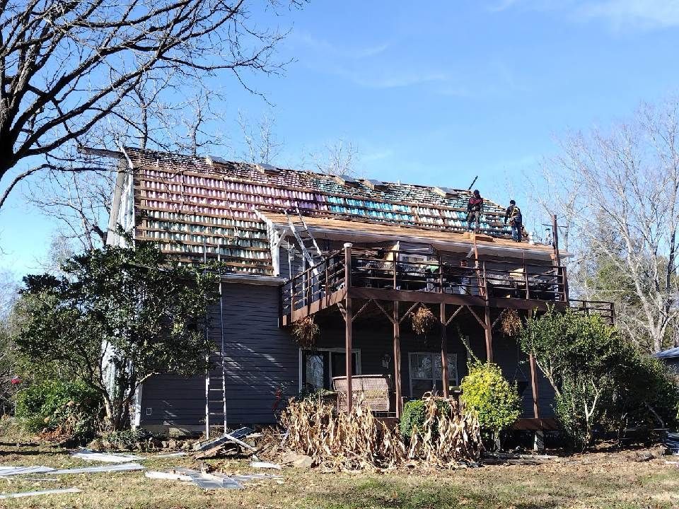 Damaged two-story house with a collapsed roof and overgrown yard under a clear blue sky