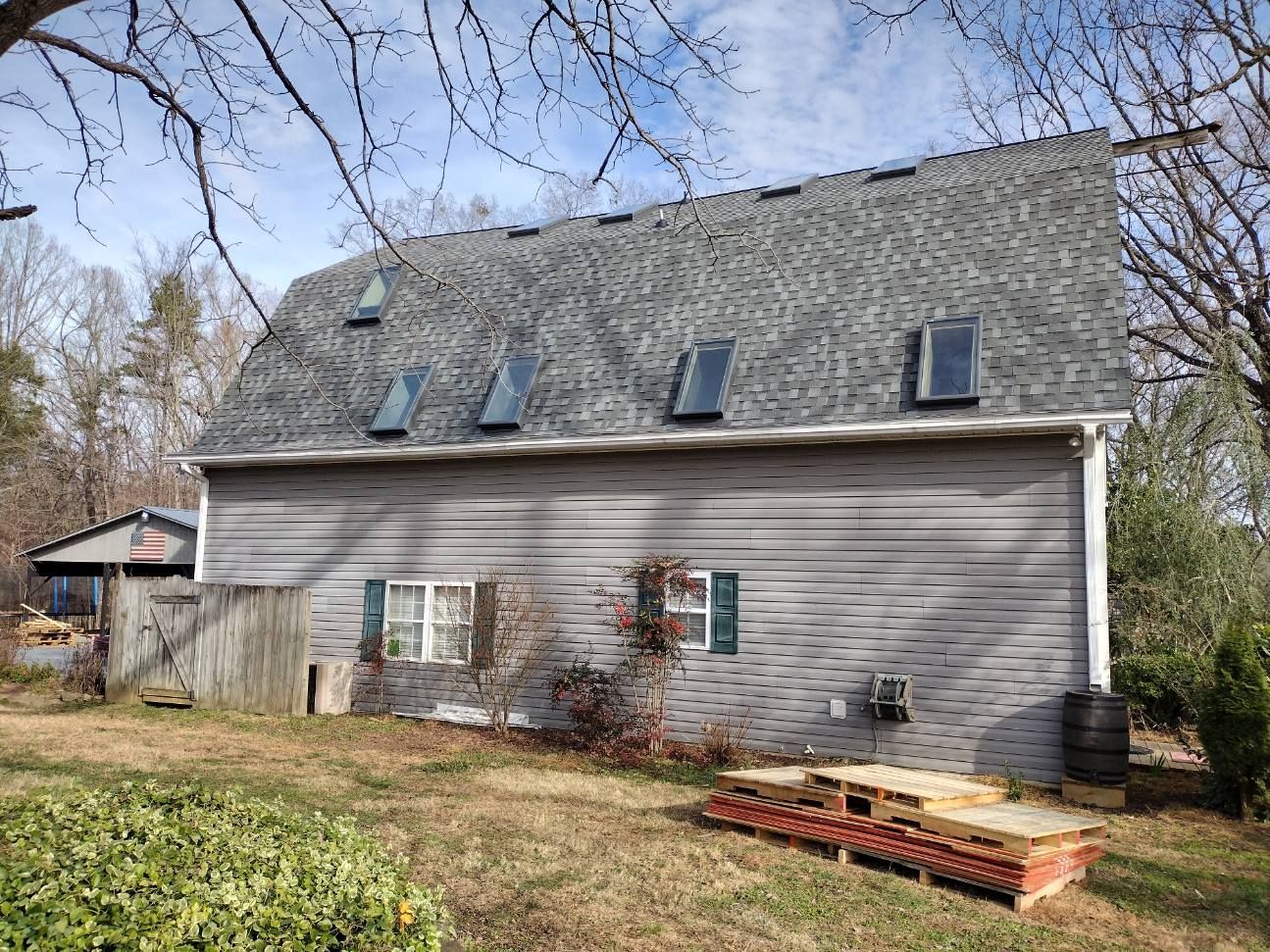 Gray house with a steep shingled roof, small windows, and a yard with bare trees and a red planter box