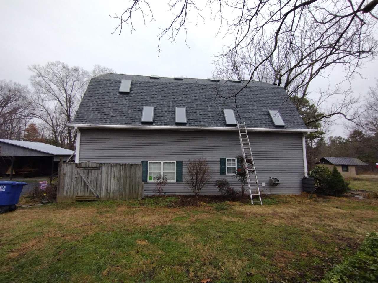 Gray house with a steep roof, small windows, and a ladder leaning against the back in a grassy yard.