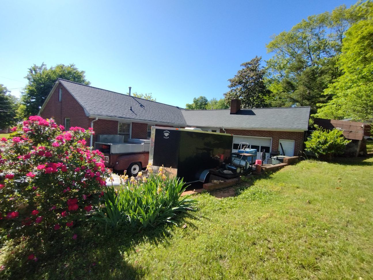 Sunlit suburban house with a carport, parked vehicles, pink flowering bushes, and a green lawn