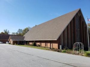 Large A-frame church building with a steep brown roof and brick walls beside a parking lot under a clear blue sky