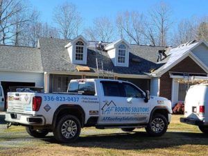 White roofing company pickup truck parked outside a house with a gray roof and dormer windows