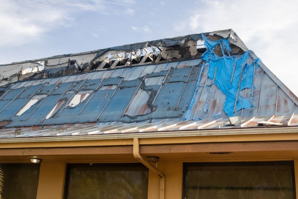 Damaged blue metal roof with torn tarps on a house, showing storm or wind damage