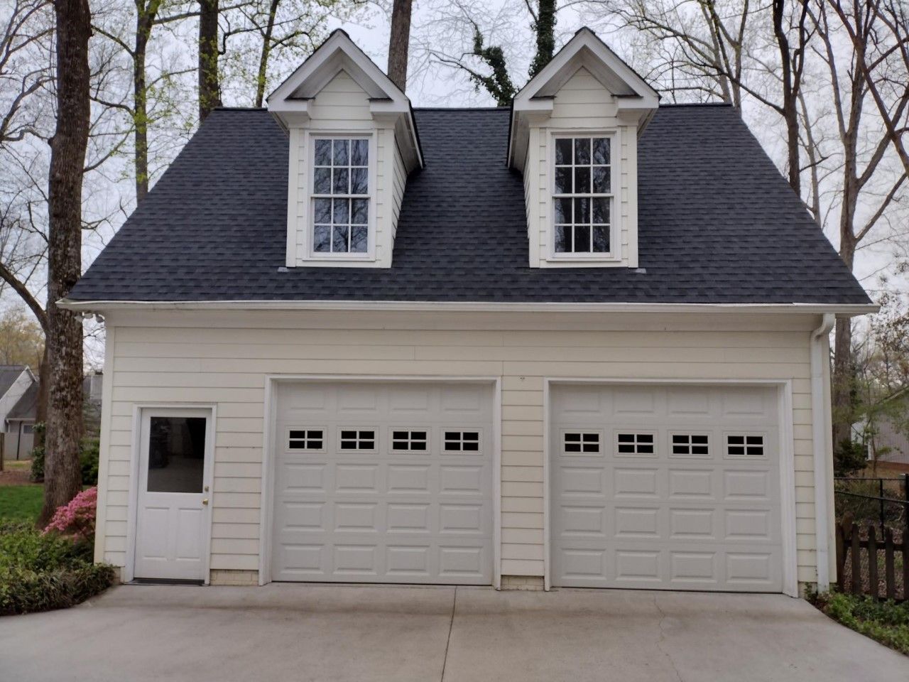 White house with two garage doors and dormer windows in a wooded setting