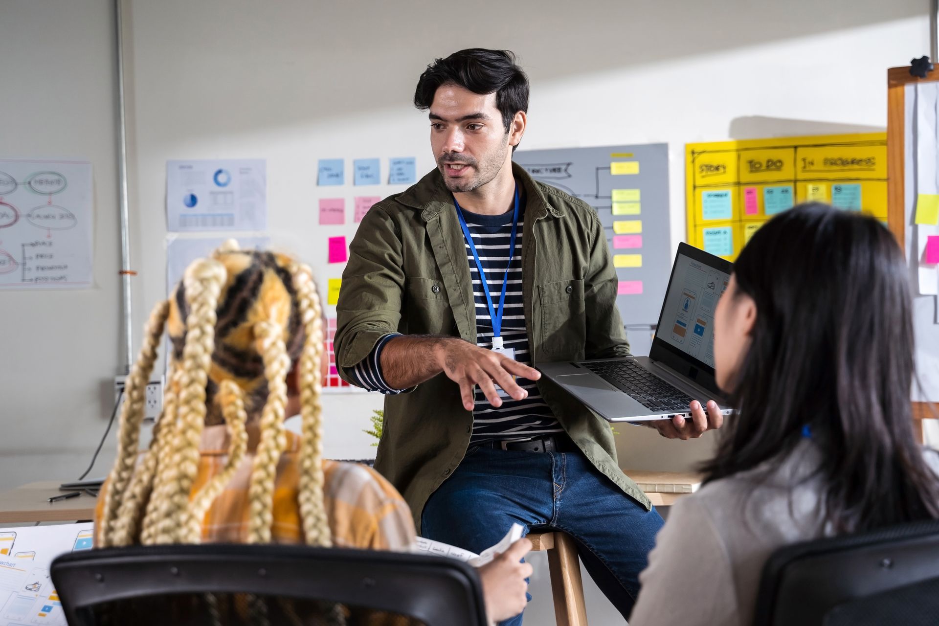 Man presenting laptop to two colleagues in an office setting; diagrams and sticky notes on wall.