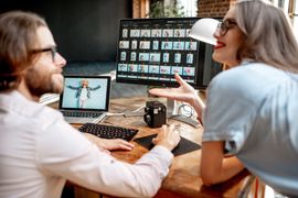 Two people reviewing photos on a computer and laptop at a desk. Woman gesturing; man using a mouse.