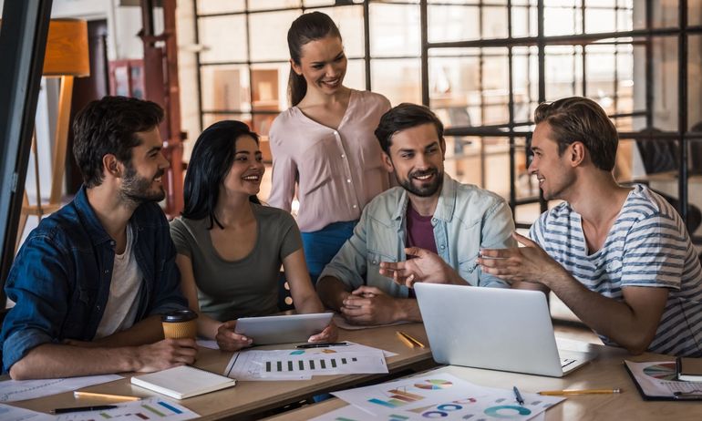 Group of people collaborating around a table with laptops, papers, and smiles in a modern office.