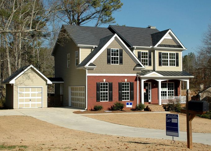 Two-story beige house with attached garage, red shutters, and a front porch on a gravel lot