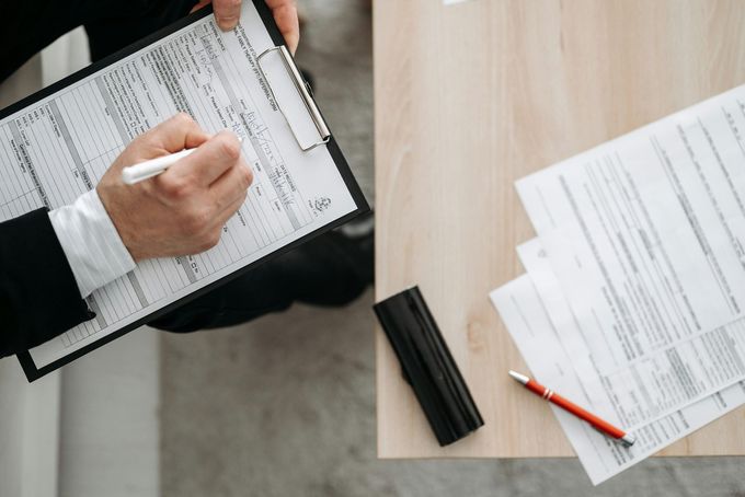 Hands writing on paperwork at a desk with scattered documents and a red pencil