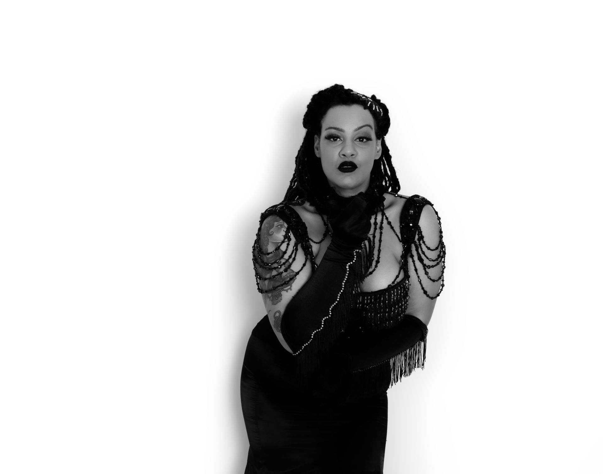 Femme en robe noire ornée de perles, gants longs et rouge à lèvres foncé, se tenant devant un fond blanc.