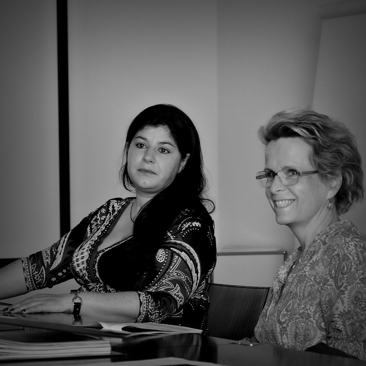 Deux femmes sont assises à une table. L'une sourit, portant des lunettes et une chemise à motifs. L'autre regarde sur le côté, vêtue d'un haut à motifs.