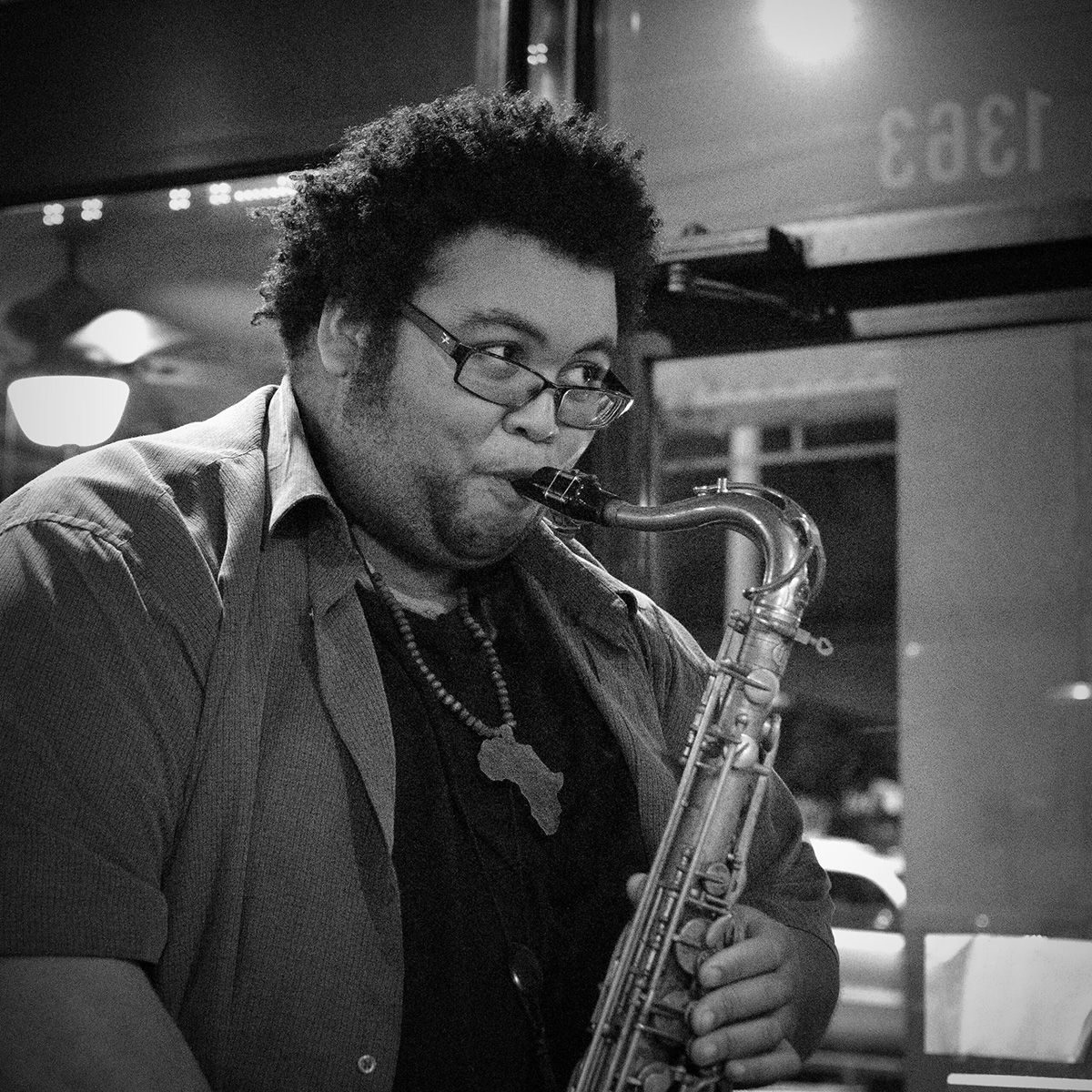 Homme jouant du saxophone, portant des lunettes, dans l'embrasure d'une porte, photo en noir et blanc.