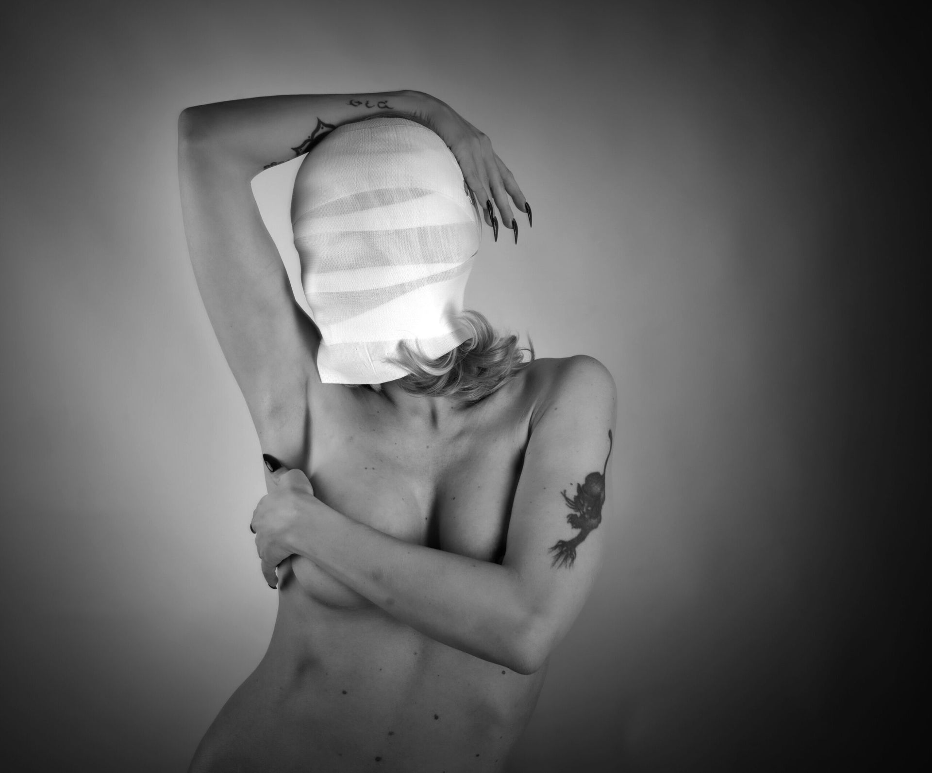 Femme la tête bandée et le bras levé, couvrant sa poitrine. Tatouages noirs et blancs sur le bras.