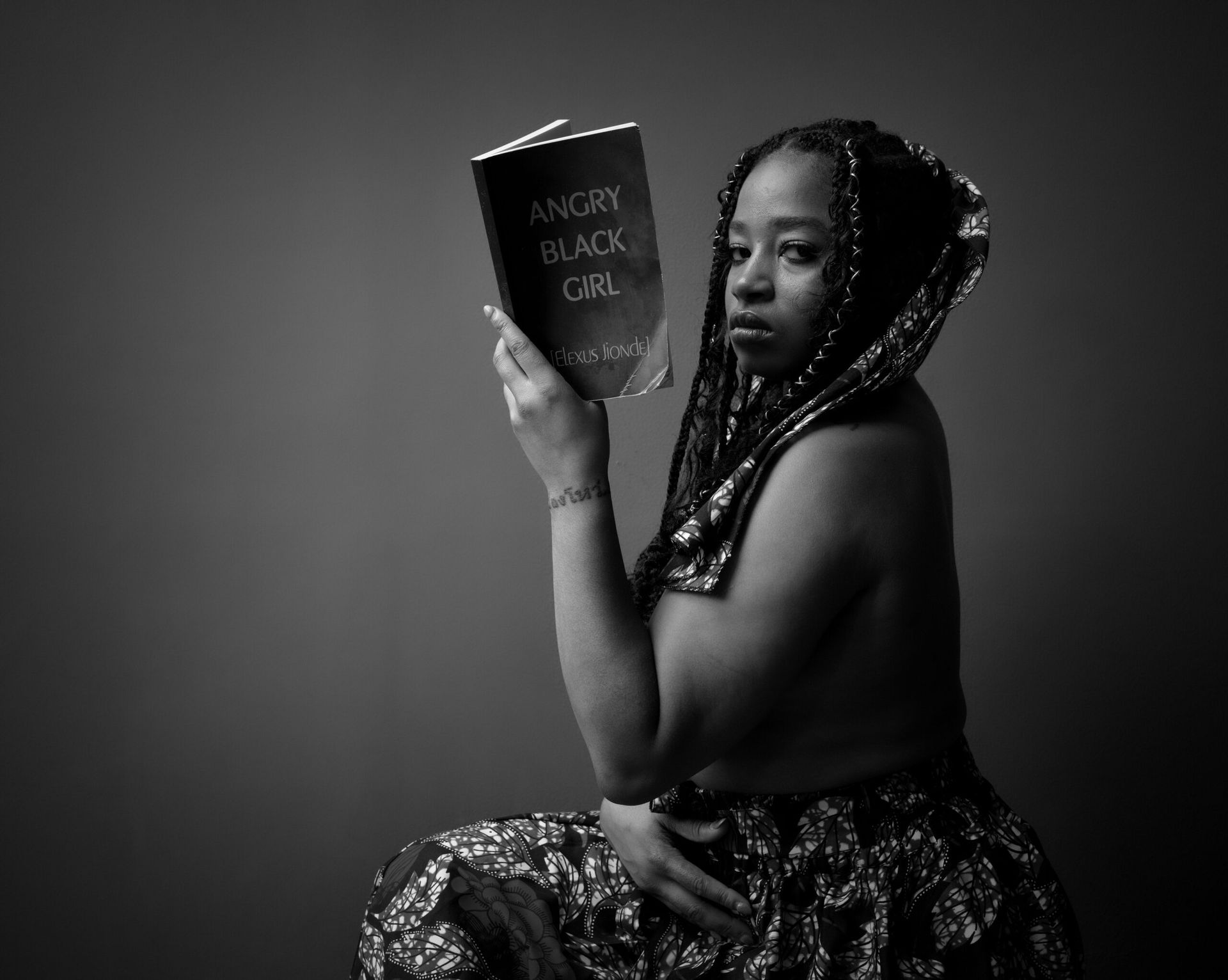 Femme vêtue d'un tissu à motifs, tenant un livre intitulé « La fille noire en colère ». Noir et blanc, assise, expression pensive.