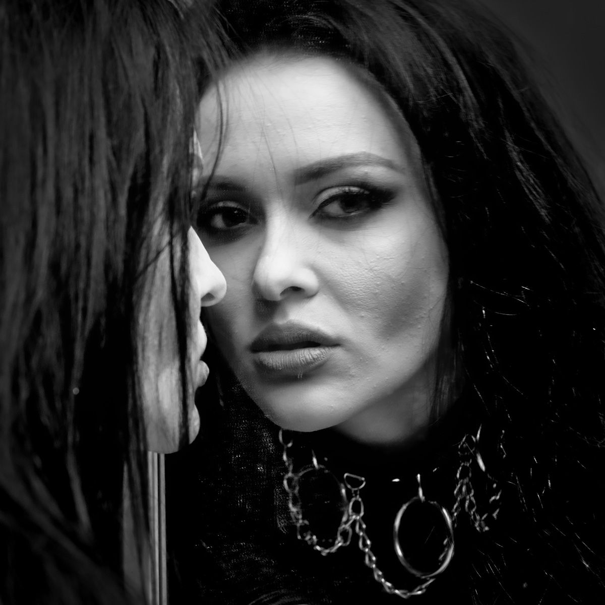 Une femme se regarde dans un miroir, portant un ras-de-cou et des vêtements sombres, en noir et blanc.