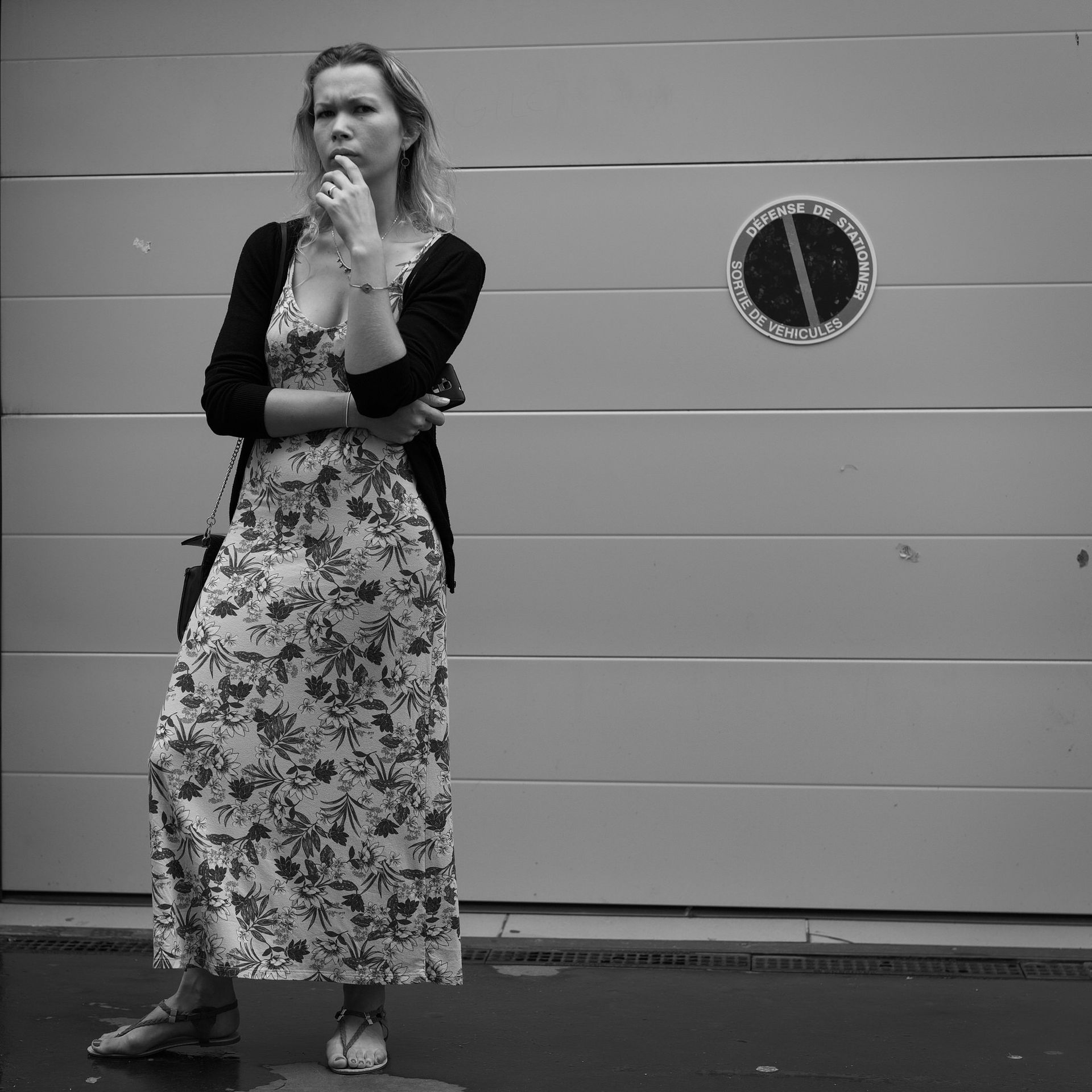Une femme en robe à fleurs, pensive, se tient debout à côté d'une porte de garage fermée avec un panneau « Stationnement interdit ».