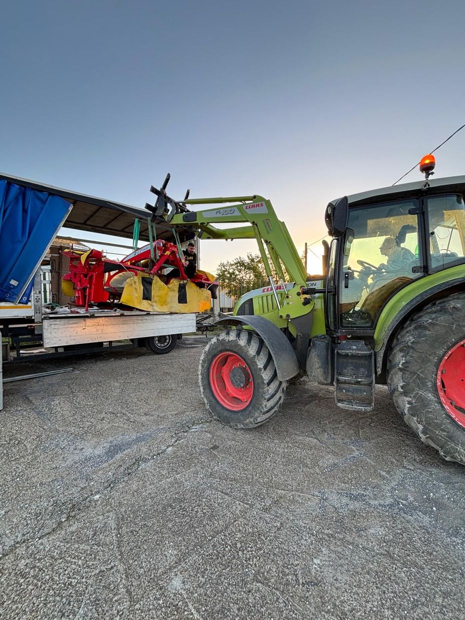 Un trattore verde carica attrezzature agricole rosse su un rimorchio al crepuscolo.