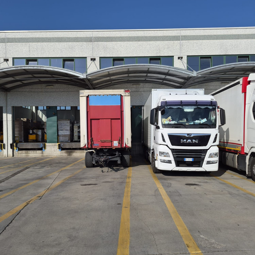 Camion parcheggiati sulla banchina di carico di un magazzino, sotto tettoie curve, tra cui un camion MAN bianco e un rimorchio rosso.