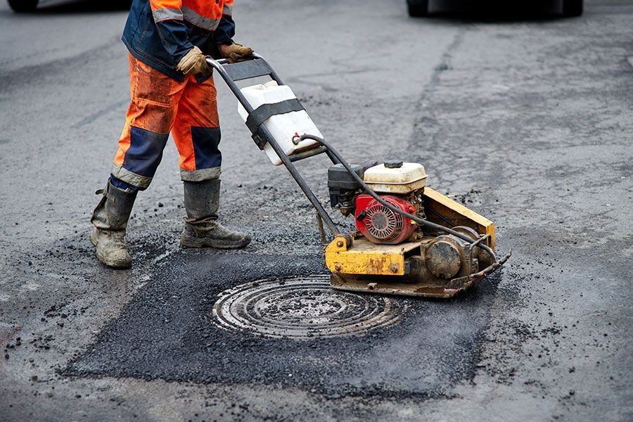 A Man Is Using a Compactor to Fix a Manhole Cover on The Road — Elite Bitumen & Asphalt In Mackay, QLD