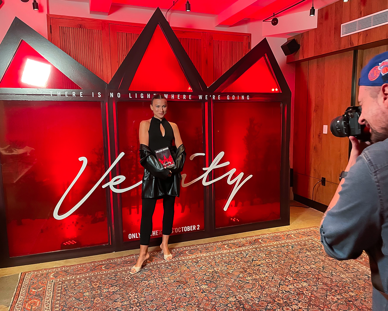 Person posing in front of a red “Verity” backdrop at an indoor event, with a photographer at right.