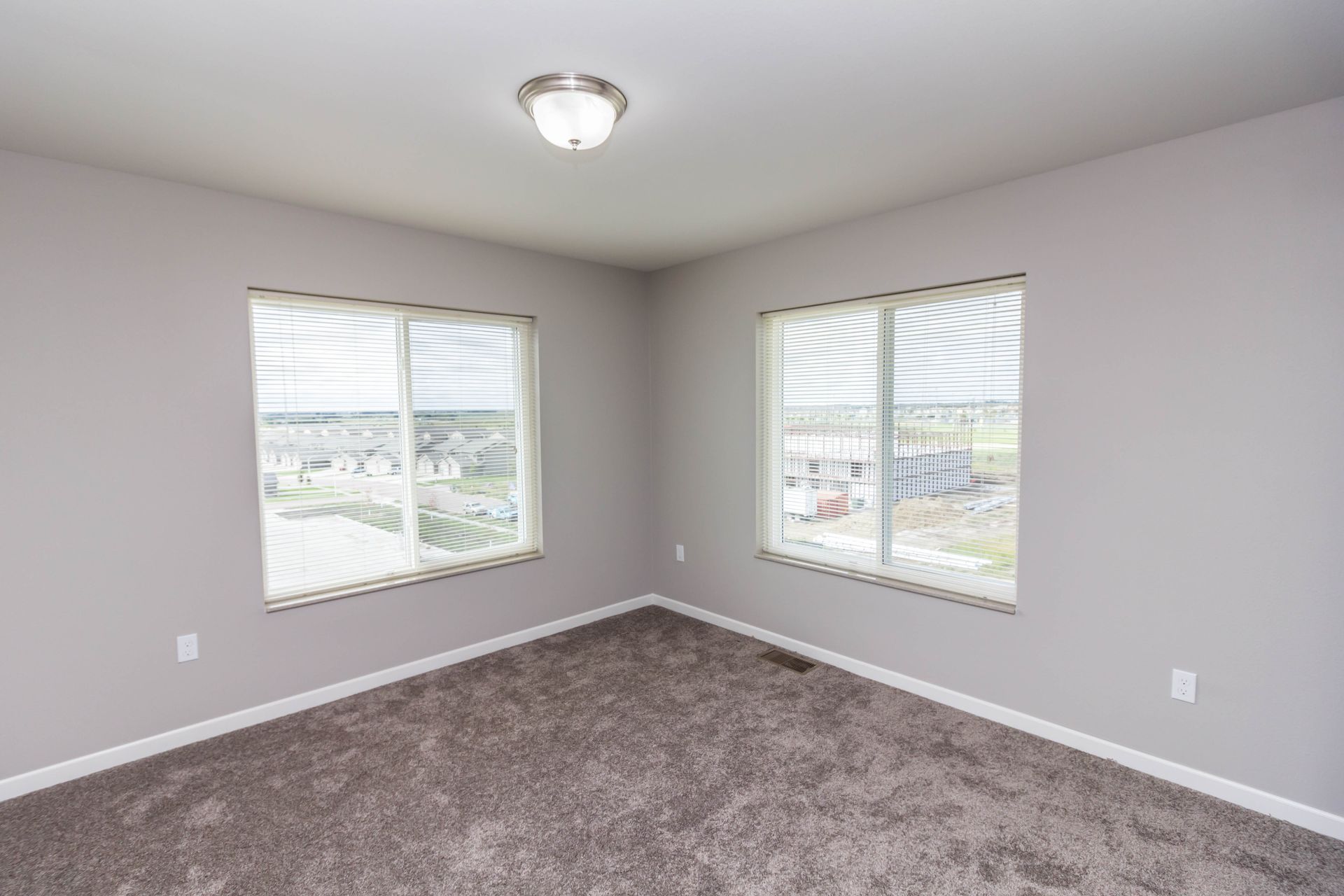 Empty bedroom with gray walls and carpet, two windows with blinds, and a ceiling light.