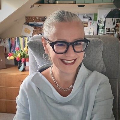 Smiling person with glasses seated in a home office, wearing a light gray hoodie and silver necklace.