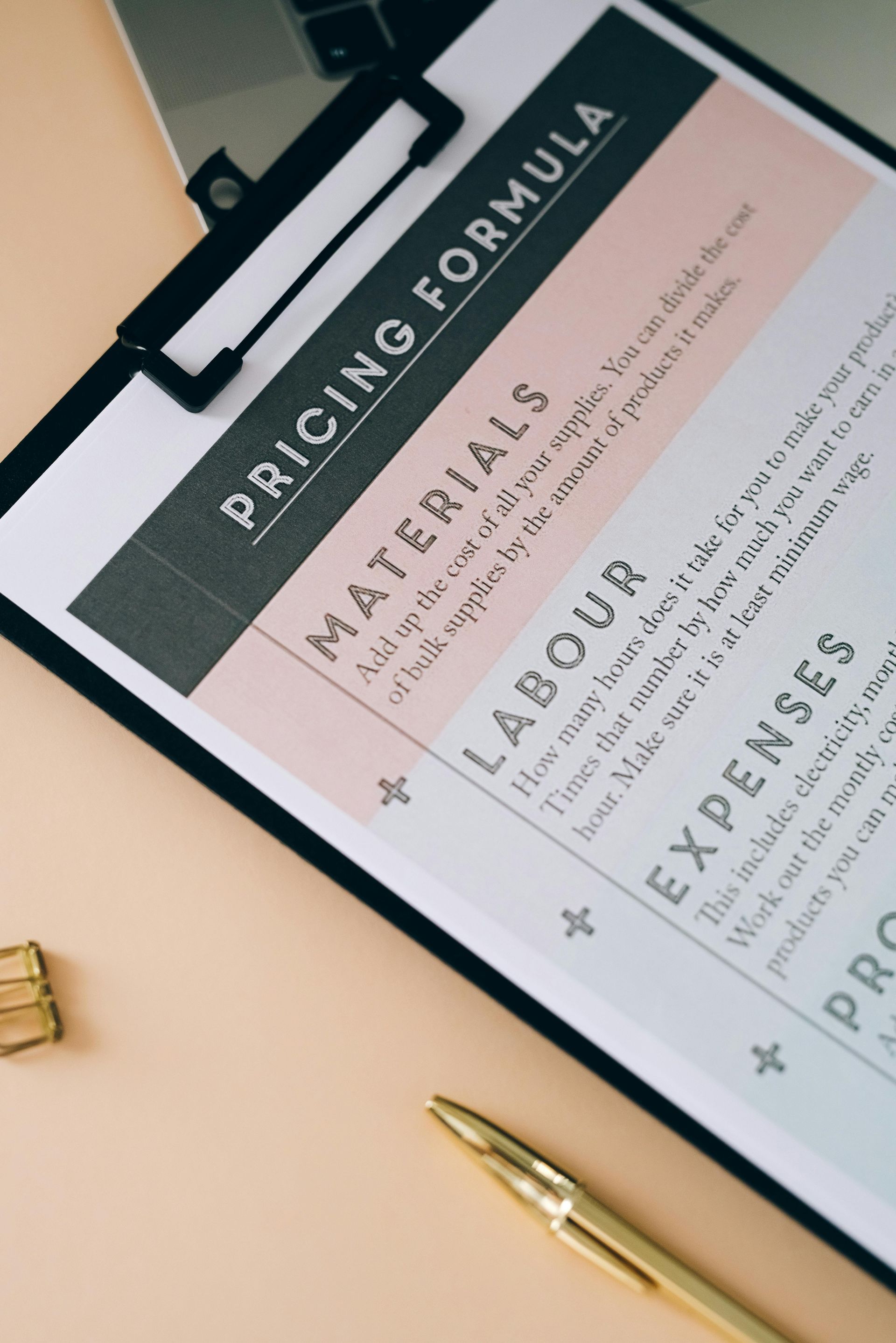 Pricing formula on a clipboard, with sections for materials, labor, expenses, and profit. A gold pen and paper clip sit nearby.