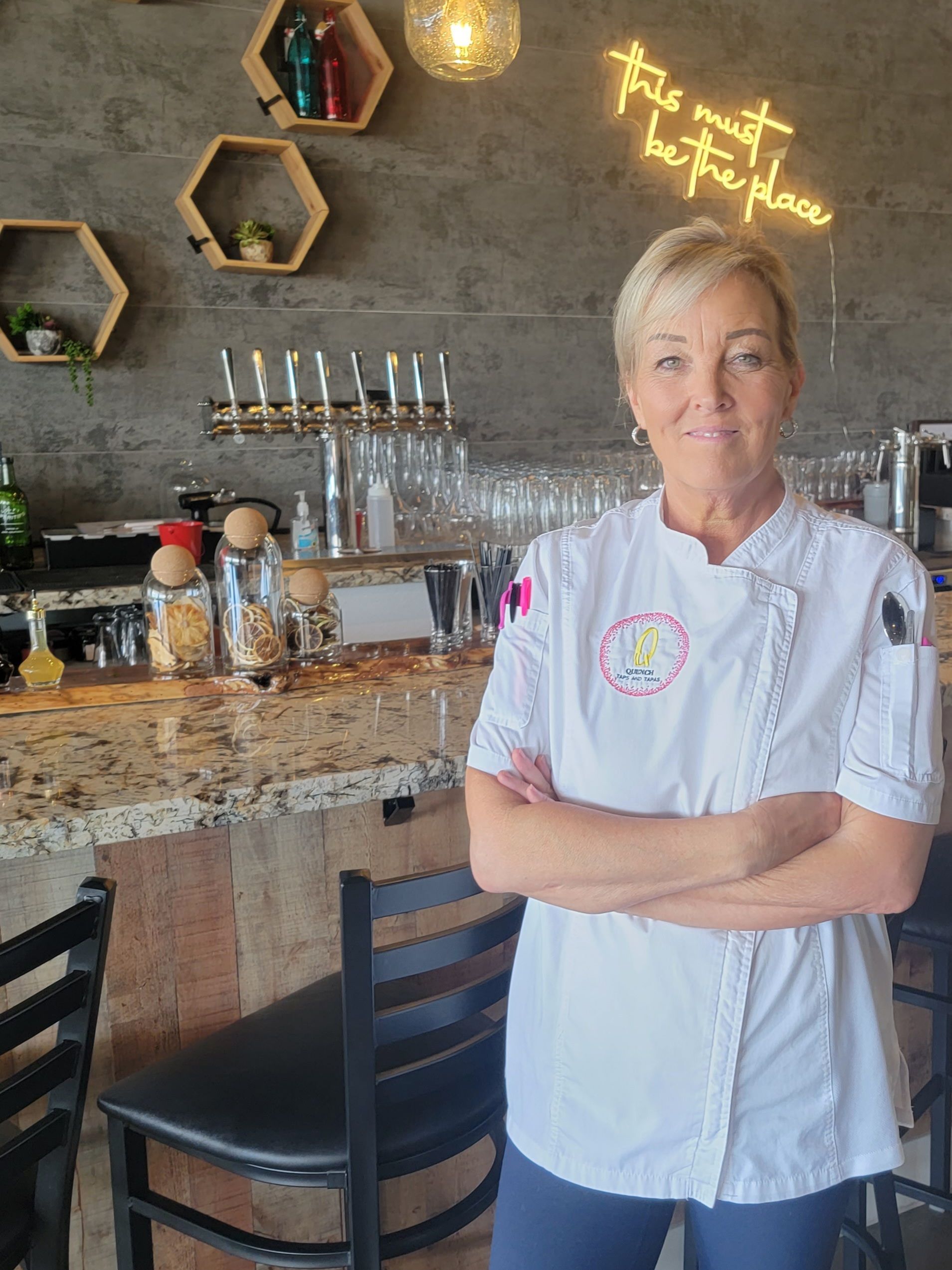 Woman in chef's coat, arms crossed, stands in restaurant. Neon sign reads