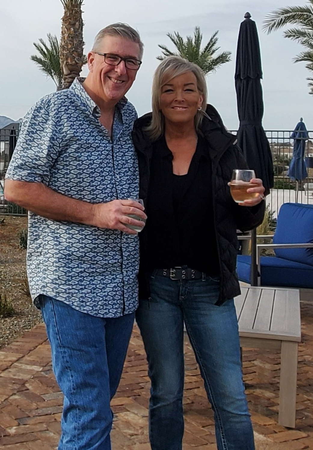 Man and woman smiling, holding drinks outside by a pool; man in blue patterned shirt, woman in black jacket.