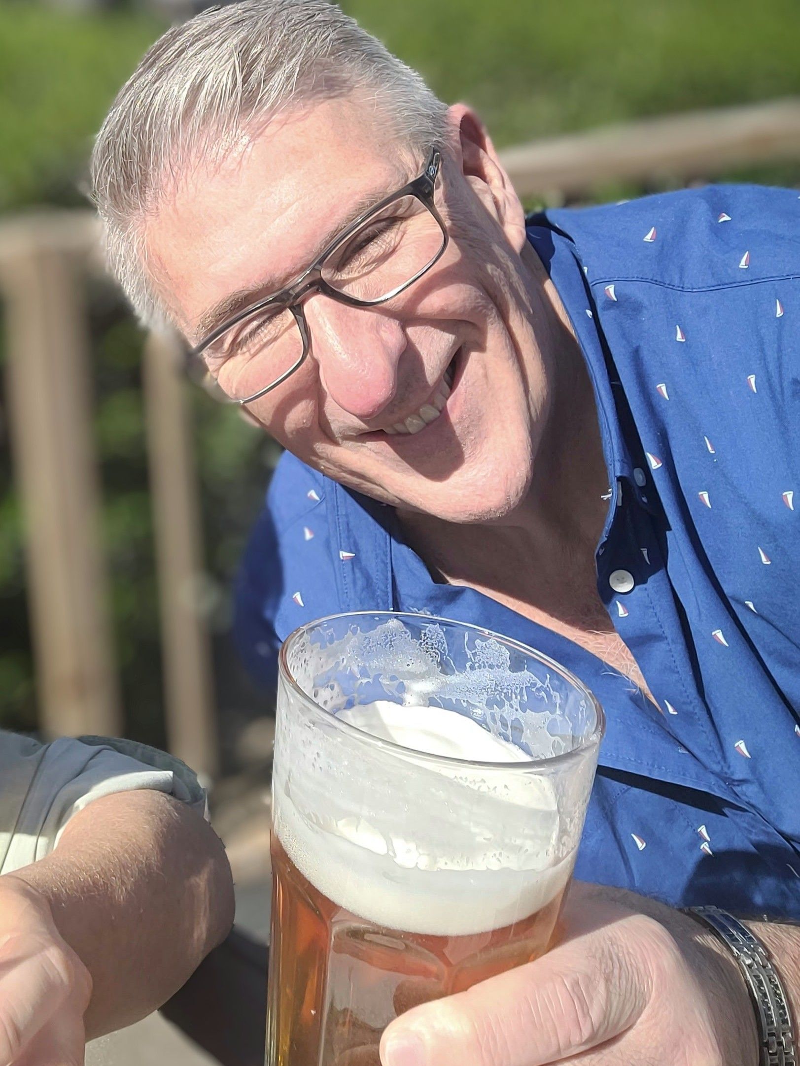 Man with glasses smiles, raising a beer glass, wearing a blue shirt, outdoors.
