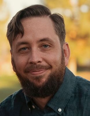 Man with brown hair and beard, smiling, wearing a blue shirt, outdoors.
