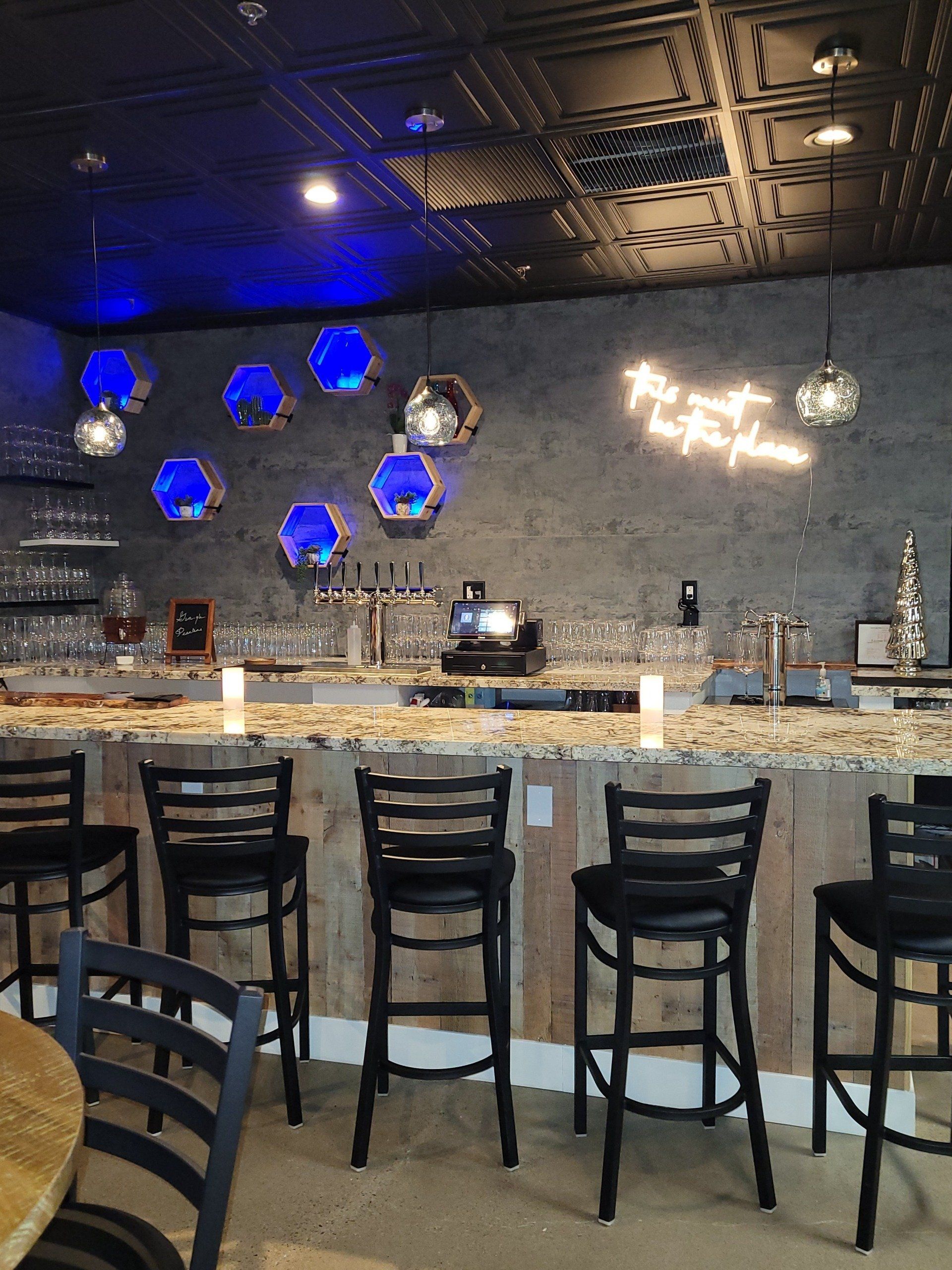 Bar with stools, granite counter, blue globe lights, neon sign on a gray wall.