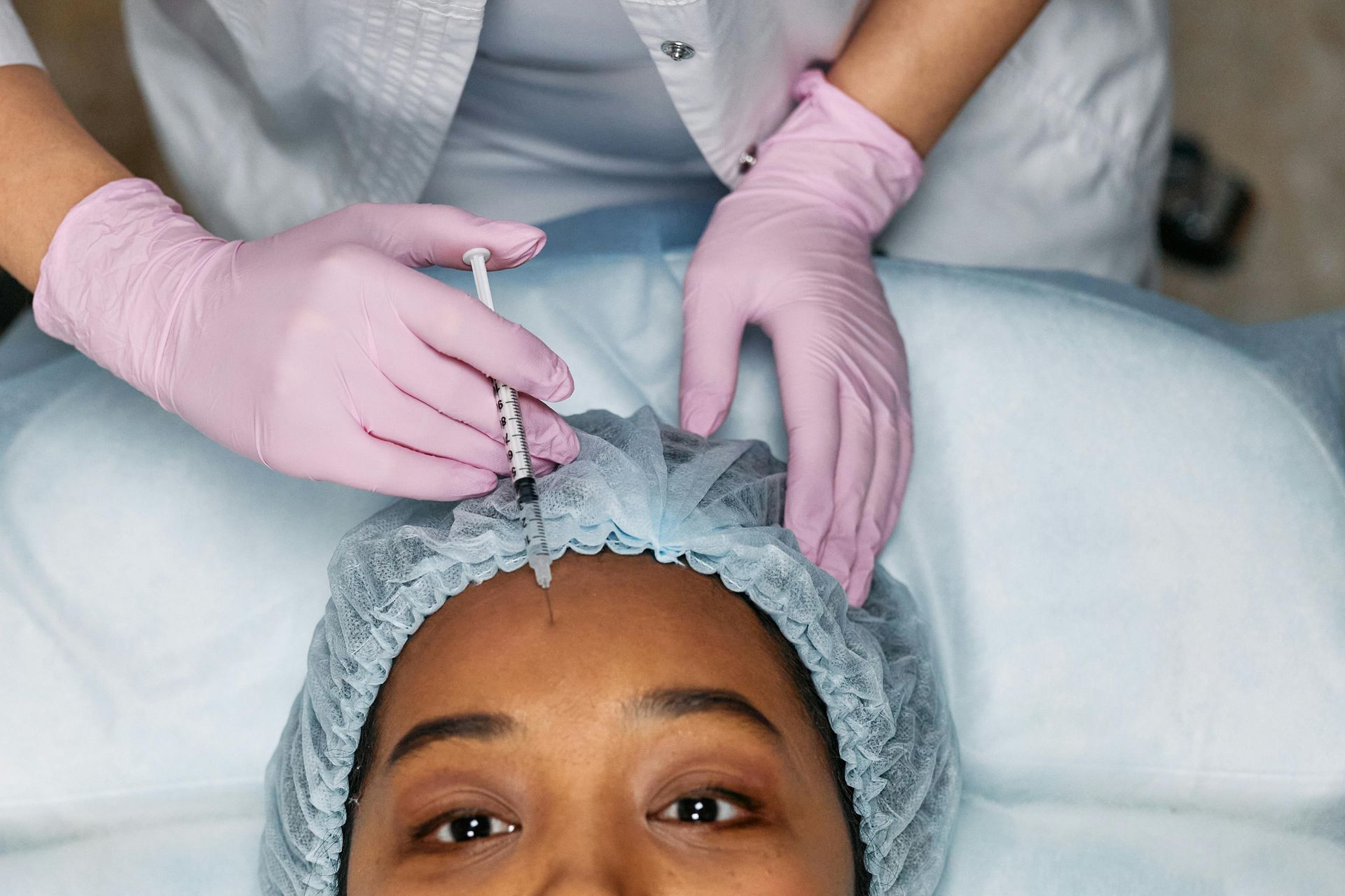 Person receiving a facial injection in forehead, by gloved hands, wearing a hair net, at a clinic.