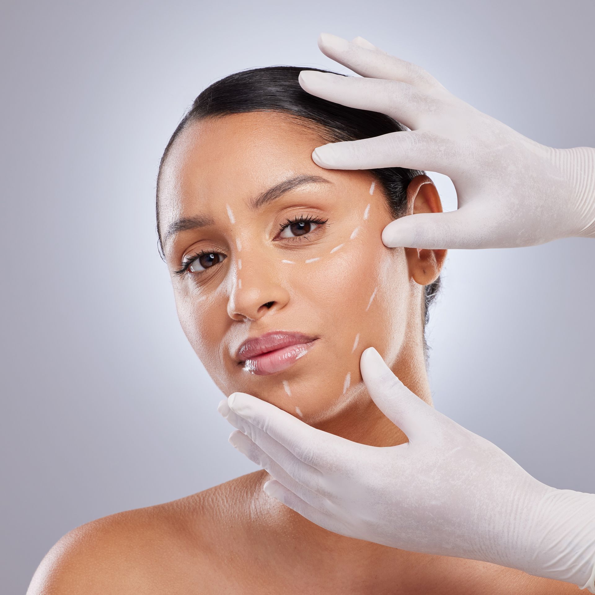 Woman with surgical markings on face; hands in gloves touch her face.