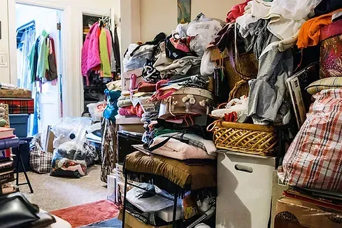 Overstuffed room with piles of clothes, boxes, and clutter. A doorway with hanging clothes is visible.