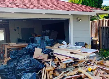 Garage exterior with piles of debris, open door revealing clutter inside, red roof, light gray siding.