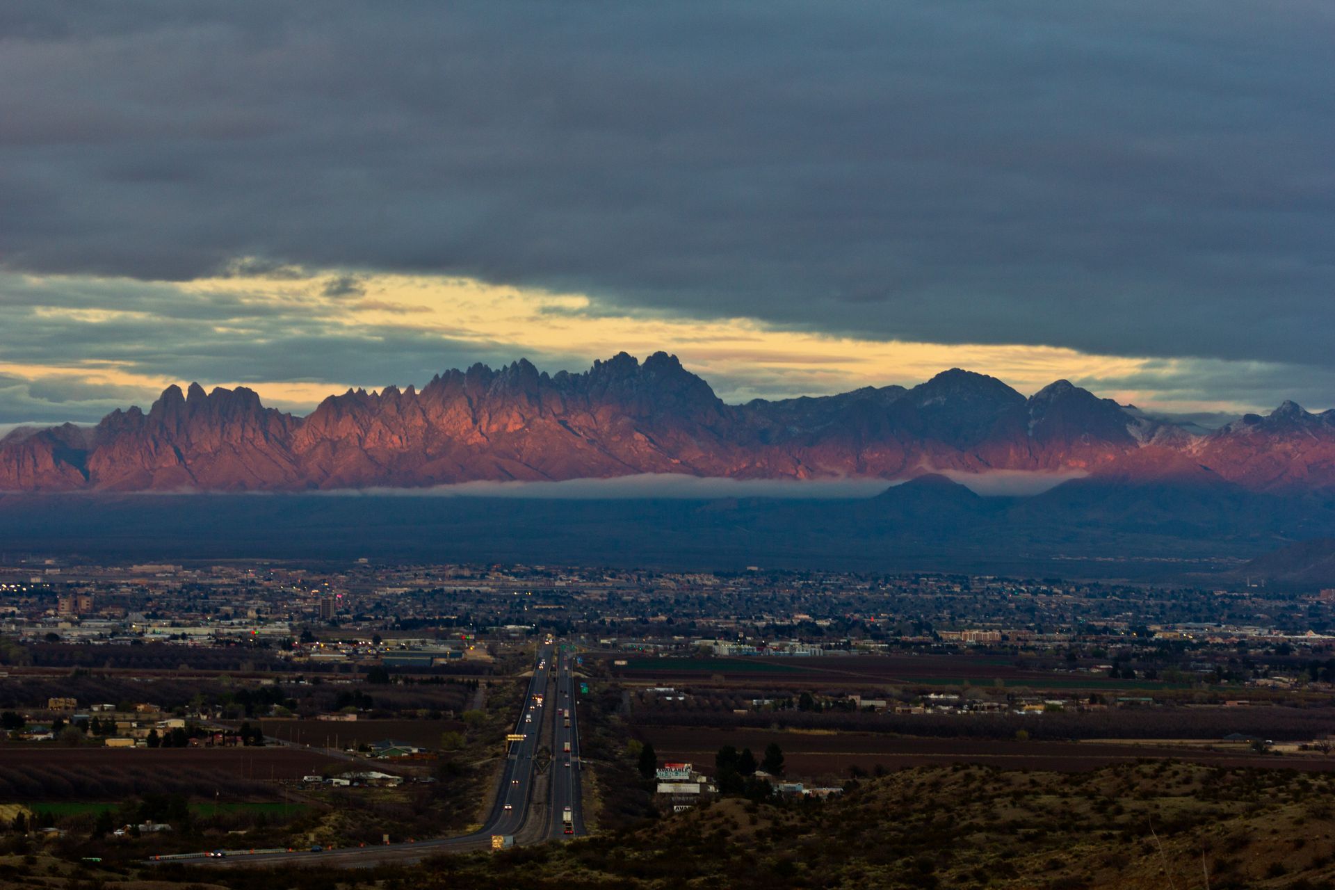Mountains lit by sunset, overlooking a city and a highway under a cloudy sky.