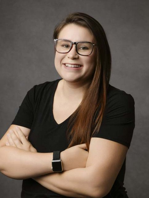 Woman with glasses and arms crossed, smiling, wearing black shirt and watch, gray background.