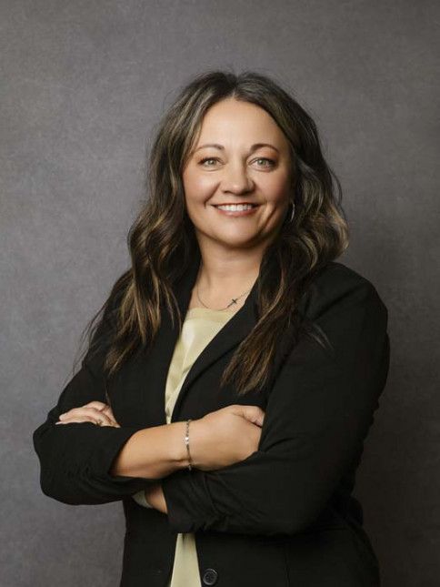 Woman with arms crossed, smiling. Wearing a black blazer and light-colored top, standing in front of a gray backdrop.