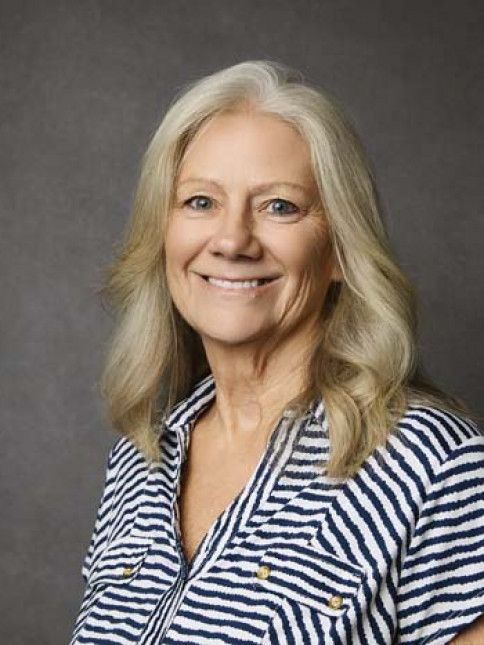 Woman with long blonde hair, smiling, wearing a blue and white striped shirt. Gray background.