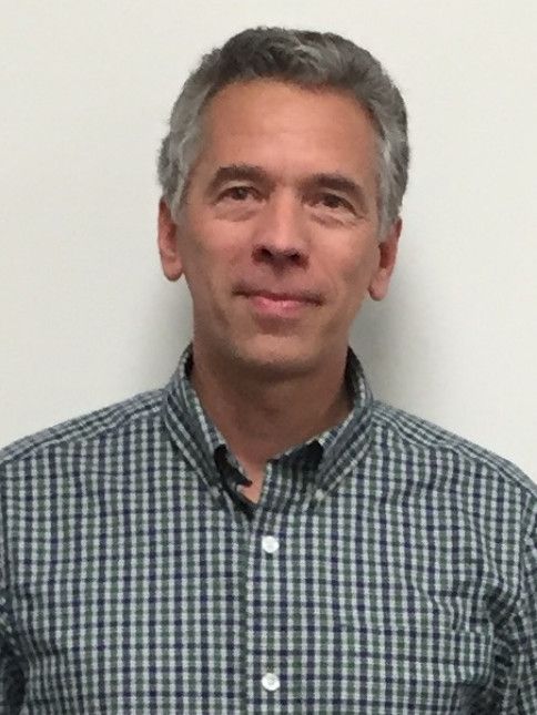 Man with graying hair in a blue and gray plaid shirt smiles slightly against a white background.