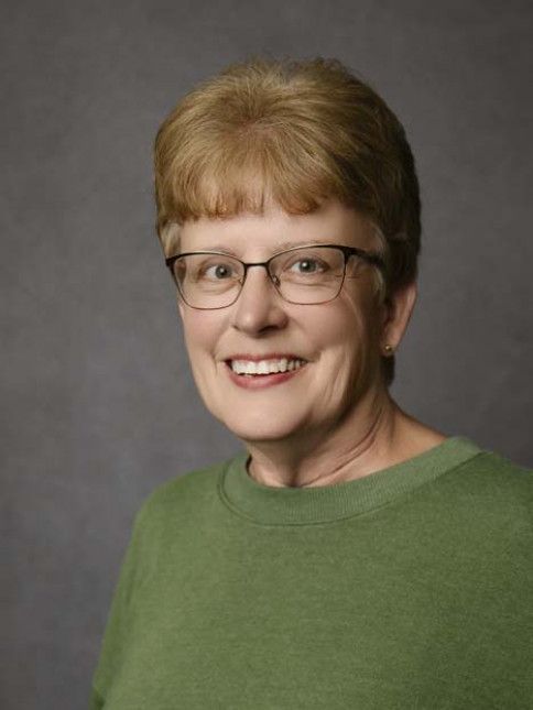 Woman with short blonde hair and glasses smiles, wearing a green shirt, against a gray background.