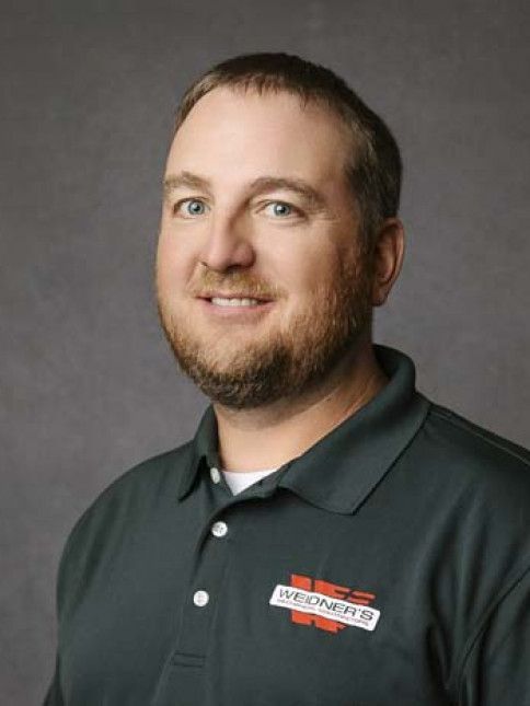 Man with a beard, wearing a green polo shirt with a logo, smiles against a gray background.