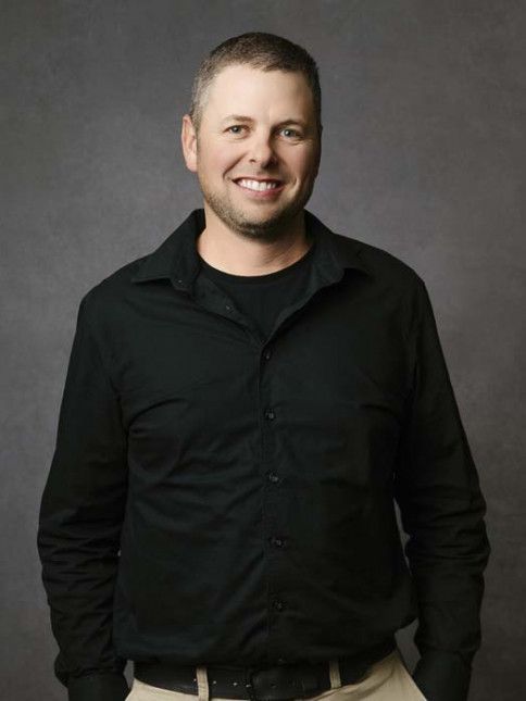 Man with short hair smiles, wearing a black button-down shirt and tan pants, posing in front of a gray backdrop.