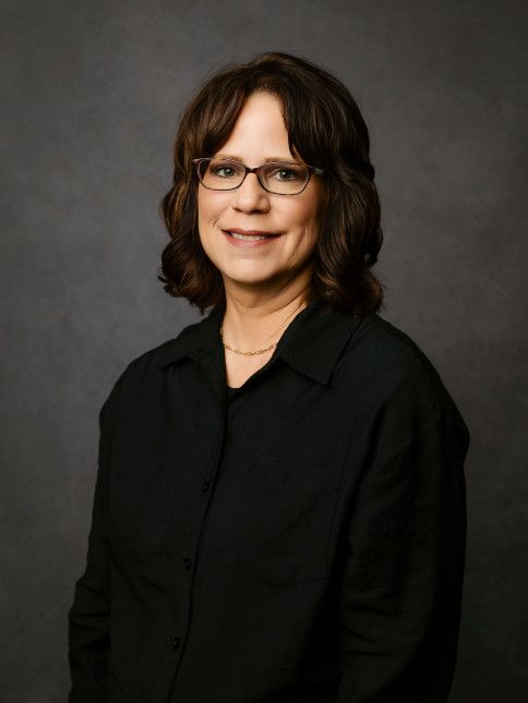 Woman wearing glasses and a black shirt, smiling against a dark gray background.