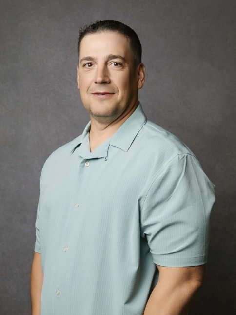 Man in a light blue button-down shirt smiles, posing against a gray backdrop.