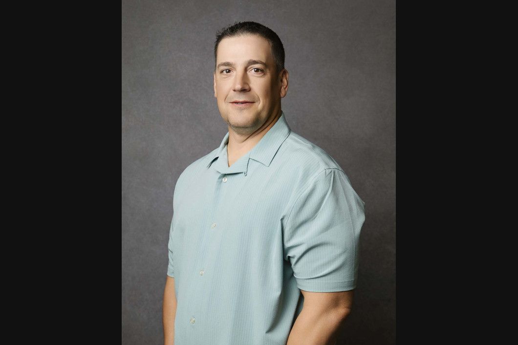 Man in light blue shirt, arms at his sides, smiling, against a gray background.